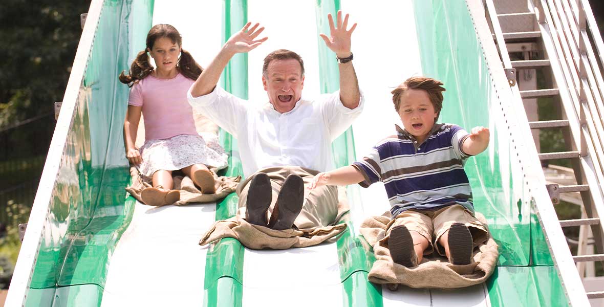 Actor and Disney Legend Robin Williams (center) glides down a large green slide with three lanes as a young girl and boy slide in the flanking lanes.