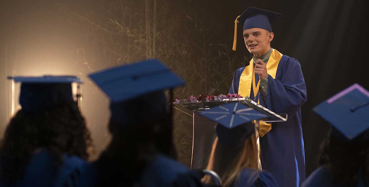 Gavin Arneson wears a blue graduation gown and matching square mortarboard with a yellow tassel hanging on the right side of it. He stands at a wooden podium and has one arm raised, giving emphasis during his speech. A group of students sit in chairs in front of him. They all wear matching gowns and mortarboards.