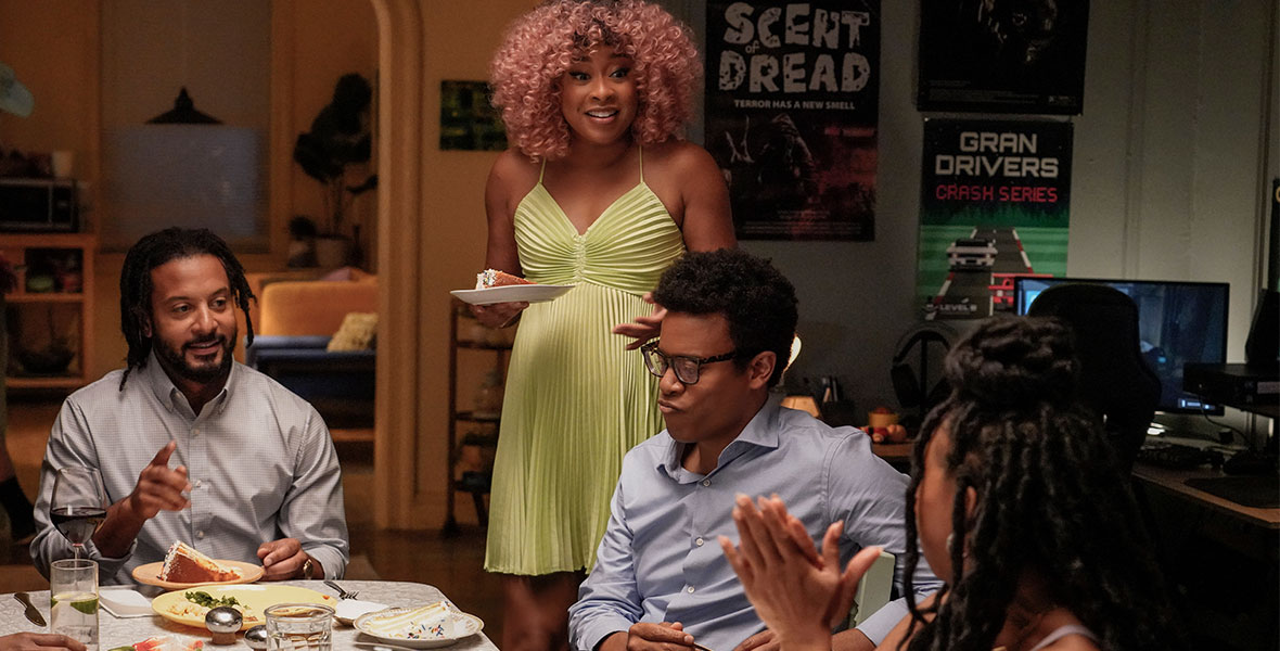 Actor Phoebe Robinson wears a neon green dress and stands next to a dining table as she speaks to her guests. Actors Jordan Carlos, Toccarra Cash, and Brandon Jay Mclaren sit around the table. The table has assorted dishes and plates in front of each guest. Behind them large movie posters and electronics are seen.