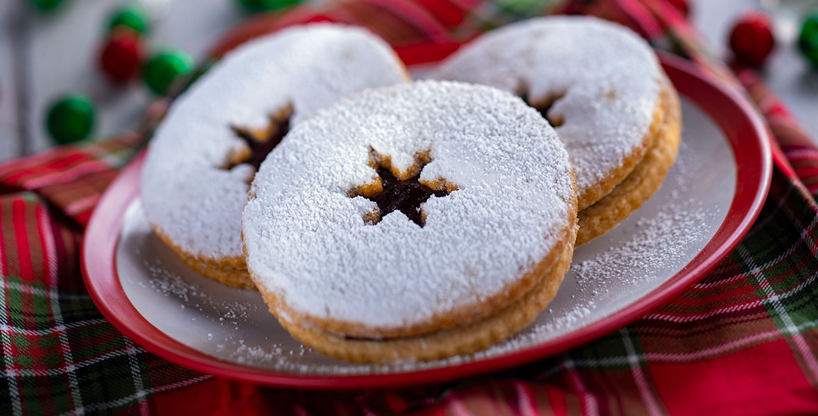 Three Linzer cookies are stacked on a white plate with a red rim. The cookies are golden and sprinkled heavily with white powdered sugar with a star-shaped cut-out in the center. The plate is lying on a red, green and white plaid tablecloth.