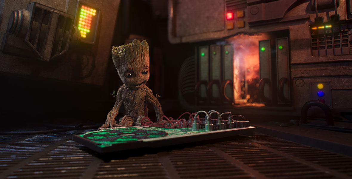 Baby Groot is sitting, smiling, and looking at a green wire board. He is inside a spaceship, and behind him are various mechanical panels, some of which are illuminated in blue, green, red, and yellow.