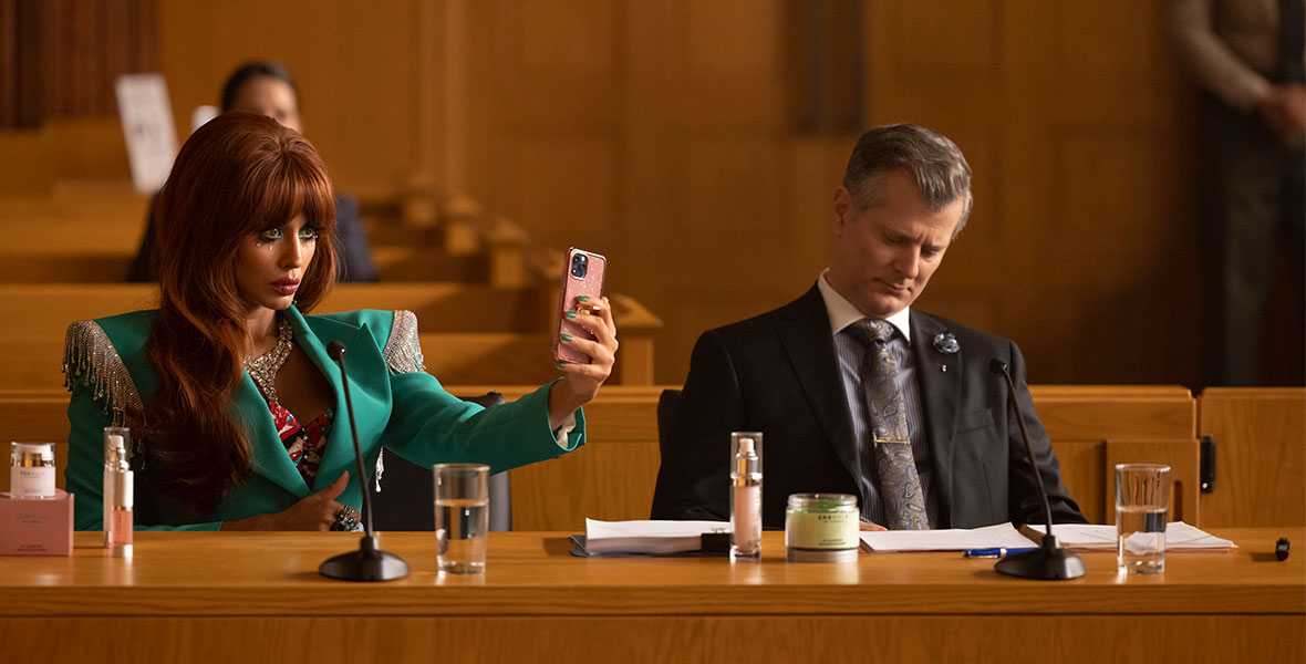 (Left to Right) Actor Jameela Jamil portraying Titania sits at a wooden table and looks at a cell phone she is holding in her left hand. Jamil wears a teal blazer adorned with silver beads and sequins on each shoulder. A male actor sits at the table to her right with empty courtroom benches seen behind them.