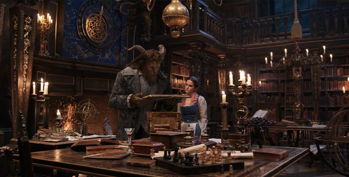 Actor Dan Stevens, portraying Beast with CGI fur and horns, and actor Emma Watson, portraying Belle, look at a book held by Stevens. Stevens and Watson stand in a large, ornate library filled with leatherbound books, chandeliers, swinging ladders, and other objects.
