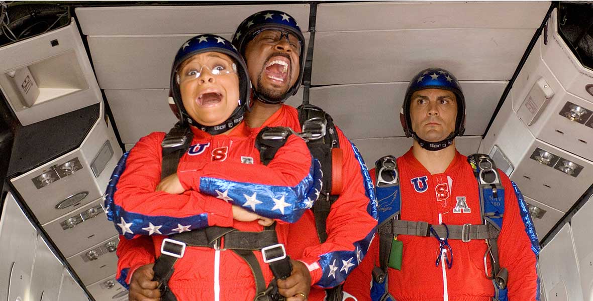 Raven-Symoné and Martin Lawrence wear red skydiving jumpsuits with blue stripes down the sleeves with white stars. Both are screaming and are strapped together. A man in a matching jumpsuit stands in the background behind them. They are all seen in the opening of a plane as they’re about to skydive.