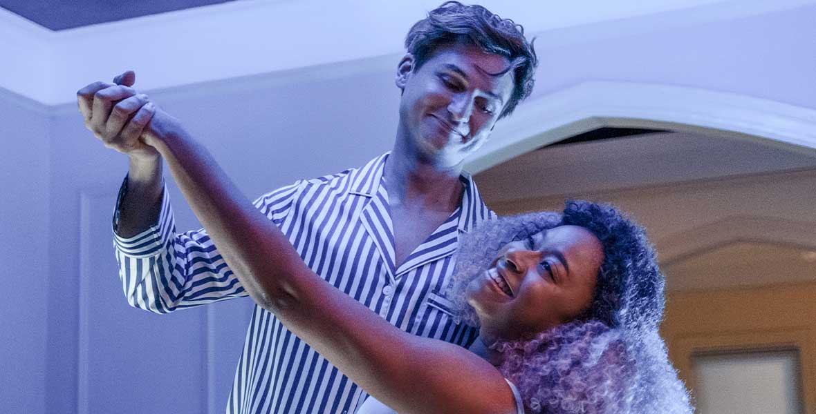 Actors Moses Storm and Phoebe Robinson ballroom dance in a dream-like sequence with soft blue lighting. Storm wears blue and white striped pajamas and Robinson wears a white tank top.