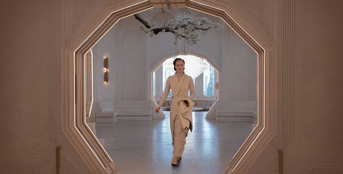 Genevieve O’Reilly as Mon Mothma, in a white robe, walks with a purpose down a white, octagonal archway. A tree with white flowers hangs from the ceiling and in the distance, a window looks out upon a city.