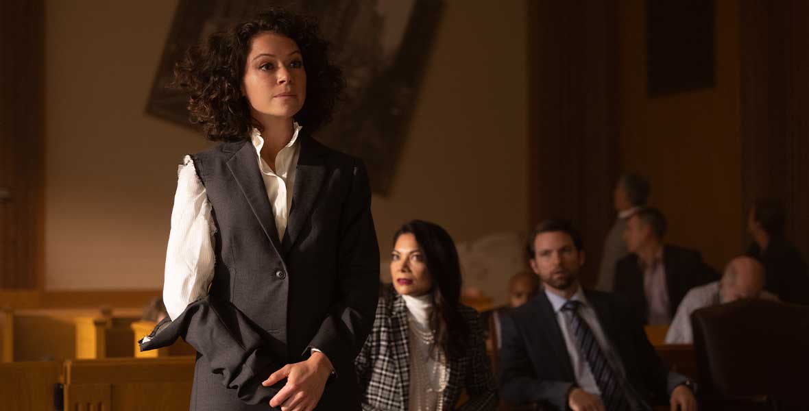 Jennifer Walters (Tatiana Maslany) stands in a court room with her hands in front of her. She is wearing a suit but the sleeve on her right arm has been ripped down to the forearm. The dress shirt is white, and her suit is dark gray. She has curly brown hair.