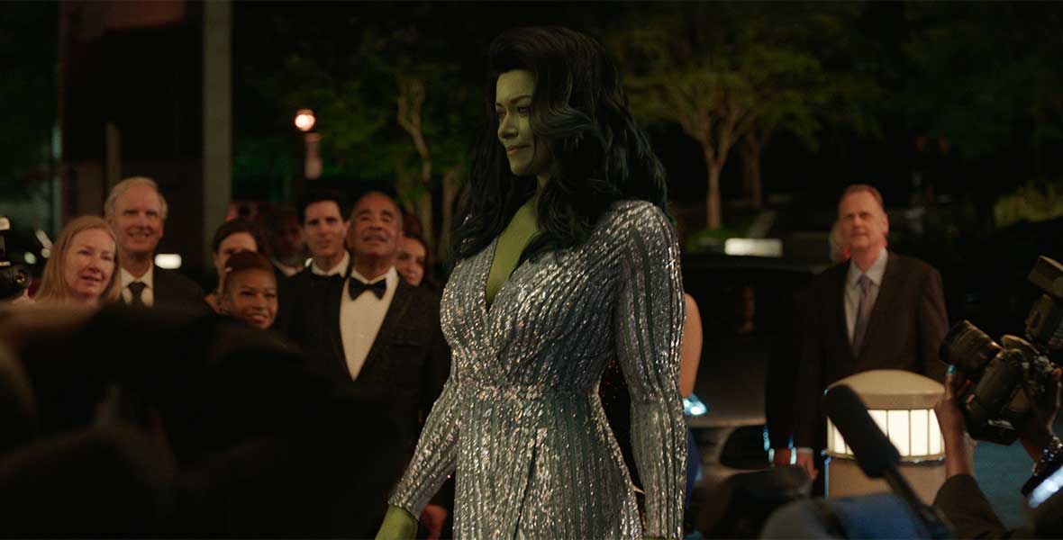 Actress Tatiana Maslany with CGI green skin and hair walks by a crowd wearing a silver beaded ballgown.