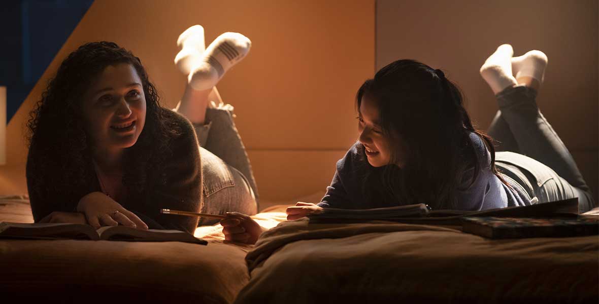 Clare Della Valle and Isabel Lam lay next to each other on their stomachs with their knees bent, elevating their legs with dark lighting. In front of each of them are books. 