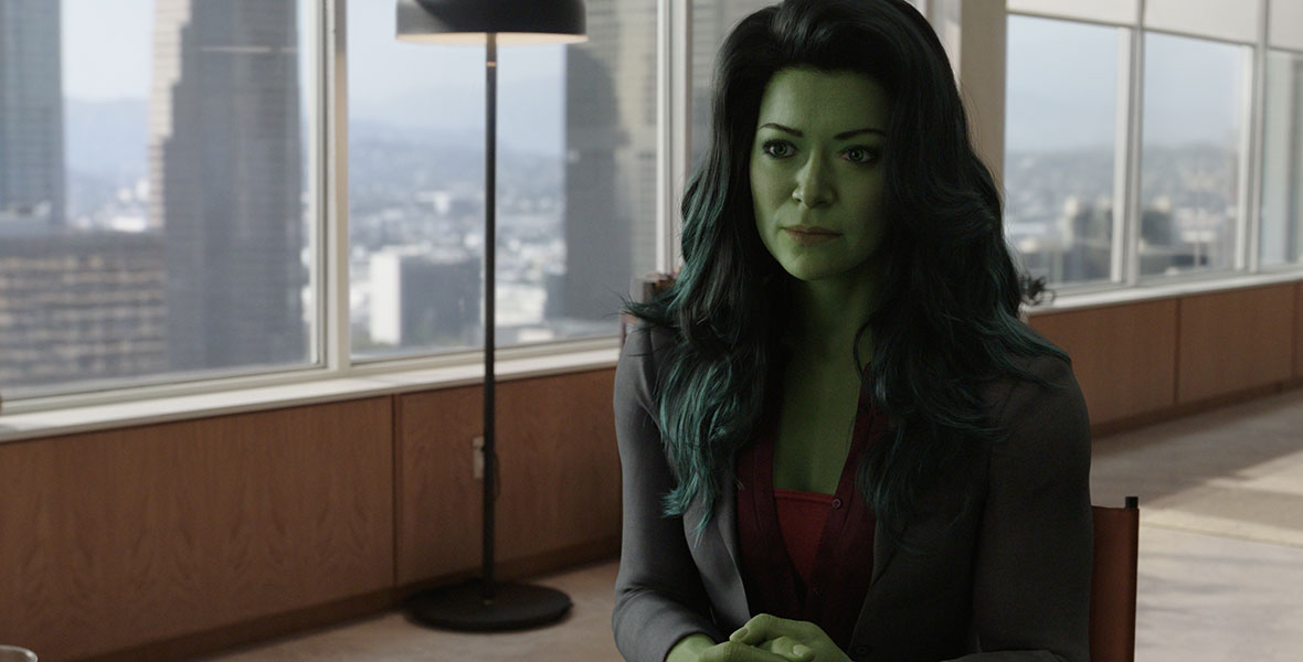 Actor Tatiana Maslany as Jennifer Walters/She-Hulk wears a navy suit jacket and sits in a chair within a large office. She has CGI green skin and hair. Behind her are large windows depicting the Downtown Los Angeles skyline.