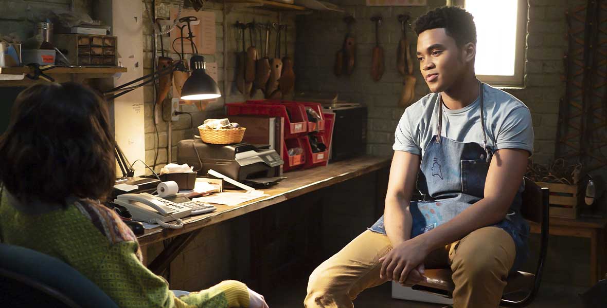 (Left to Right) Actors Devyn Nekoda and Chosen Jacobs sit across from each other in a dimly lit back room with office supplies and shoe-making materials in the background