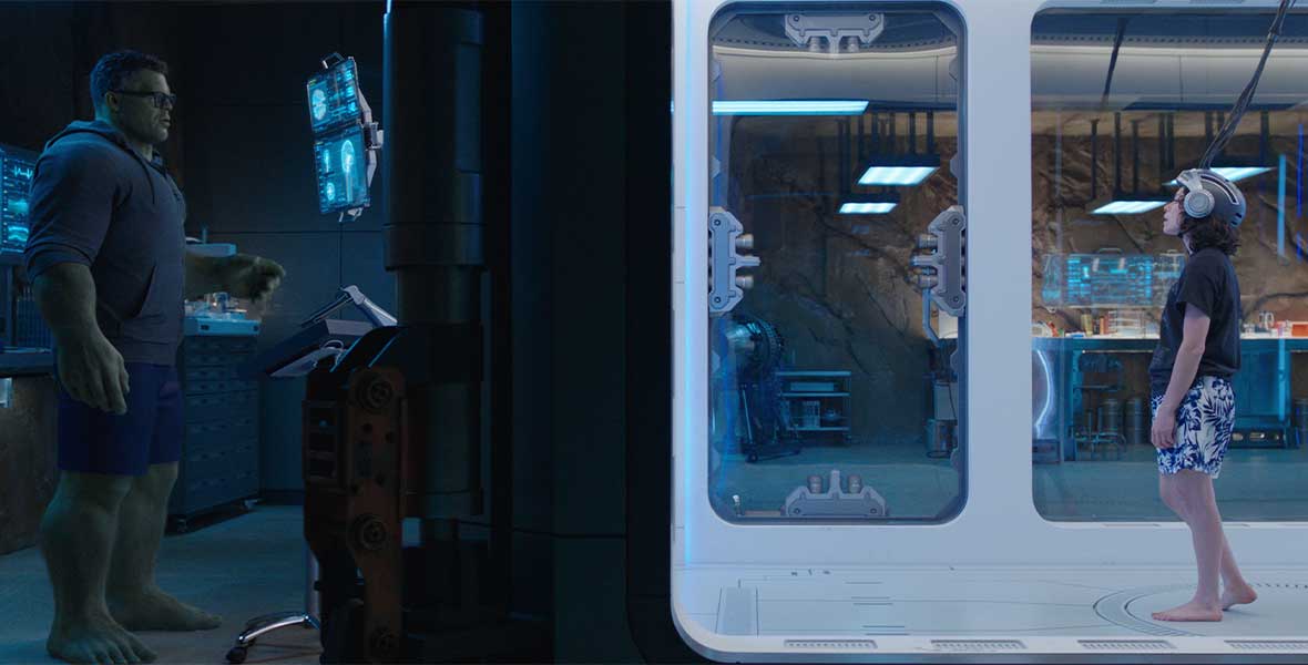 (Left to Right) Actor Mark Ruffalo as Bruce Banner with CGI green skin and hair stands outside of a large bunker filled with large machines. Actor Tatiana Maslany as Jennifer Walters stands inside the cube wearing a helmet while Ruffalo monitors her transformation into She-Hulk.