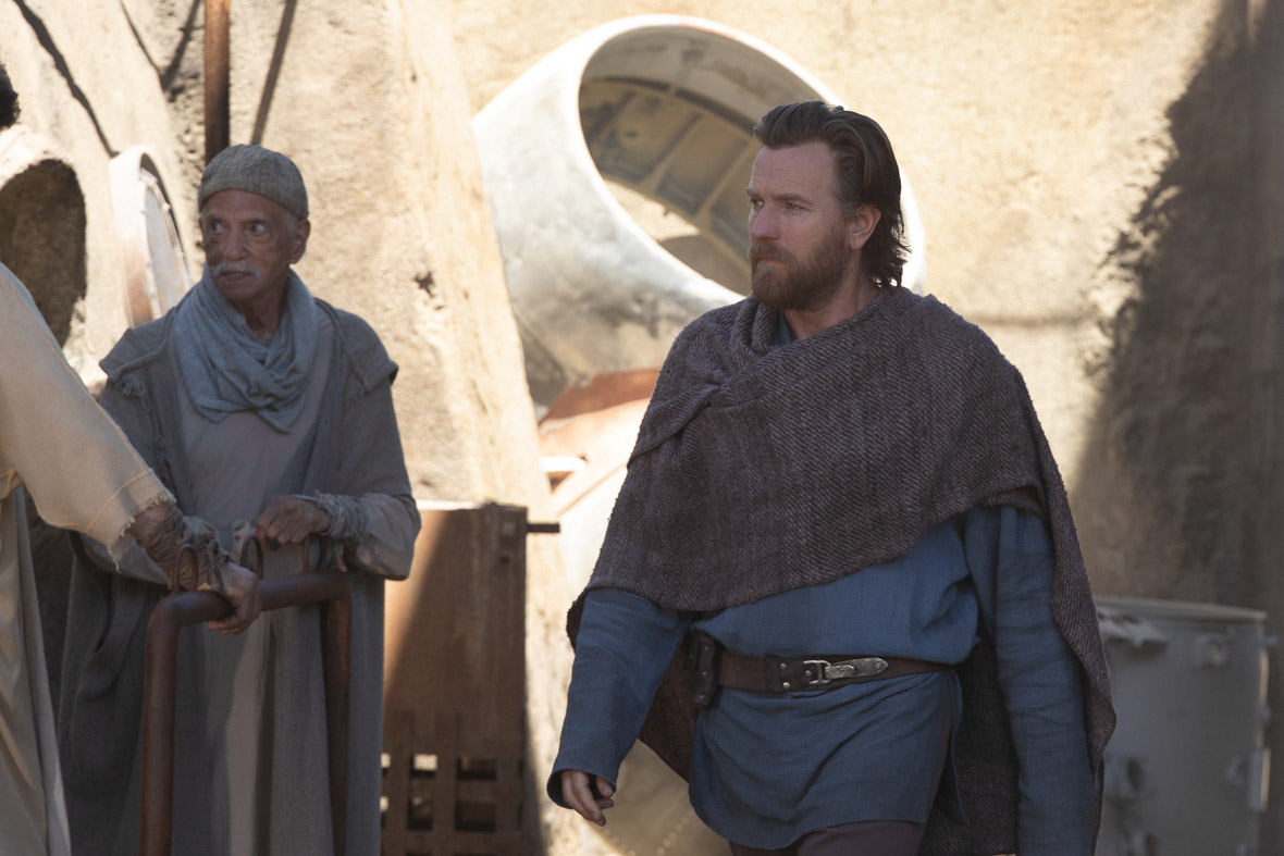 bi-Wan Kenobi, played by Ewan McGregor, walks through town. He is wearing a blue shirt, a brown leather belt, and a scarf.