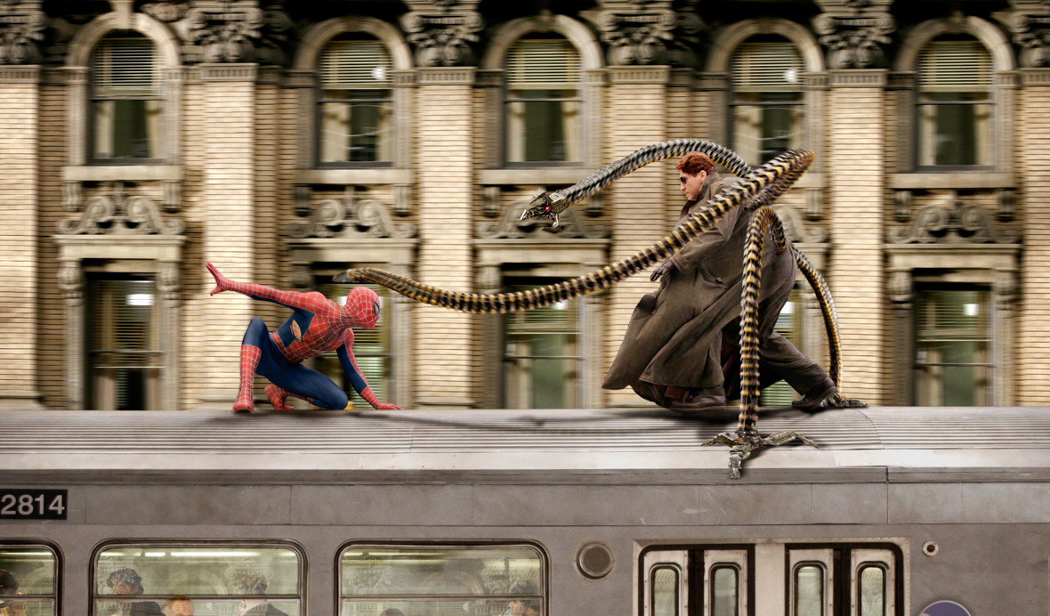 Spider-Man (Tobey Maguire) is crouched down on a train as the electronic octopus arms of Doctor Otto Octavius (Alfred Molina) reach out to attack him. There is an older beige building in the background.