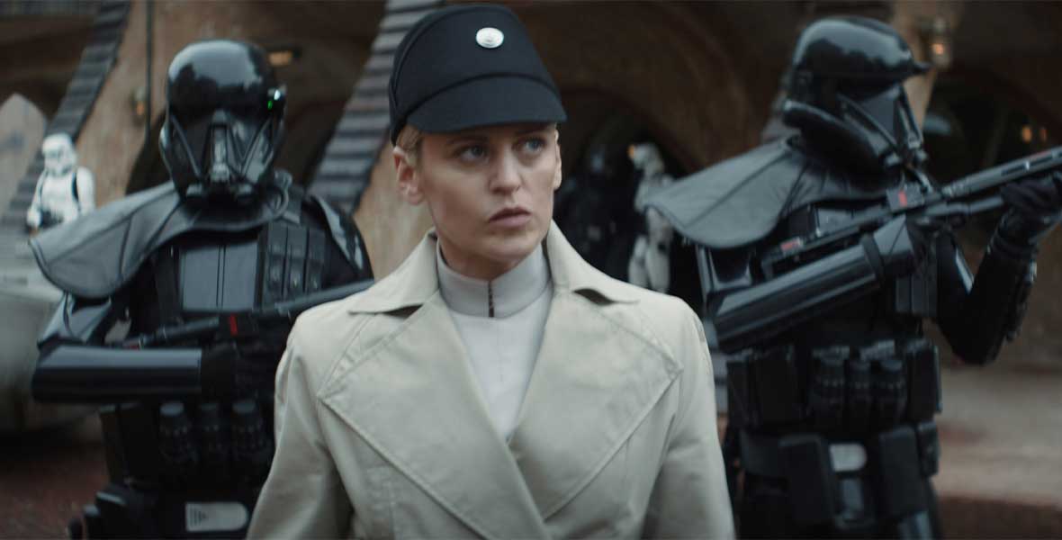 Denise Gough as Dedra Meero walks through a street in a black, Imperial cap and a white coat, flanked by two Death troopers.