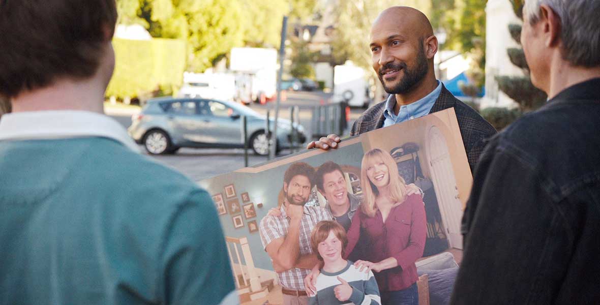 In a still from Hulu’s new series Reboot, Keegan-Michael Key is holding a photo of the cast of his former sitcom, which features him alongside Johnny Knoxville, Judy Greer, and Calum Worthy as their characters all stand in the sitcom’s living room set. He’s showing the photo to two people who have their backs to the camera, and they’re standing in a parking lot.