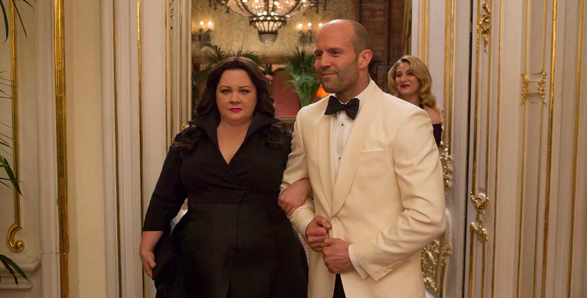 Actress Melissa McCarthy and actor Jason Statham link arms while wearing formal attire as they walk through a hallway.