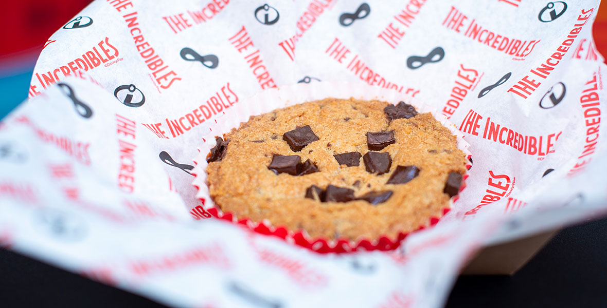 The cookie has large chocolate chunks and is baked in a red liner. The cookie sits in a basket lined with white parchment paper, reading “The incredibles” in red writing. 