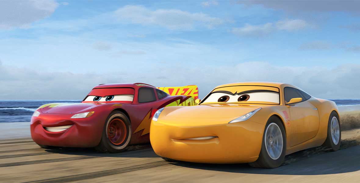Animated cars Lightning McQueen and Cruz Ramirez race on a sandy beach with a blue sky behind them. McQueen is a bright red sports car and Ramirez is yellow.