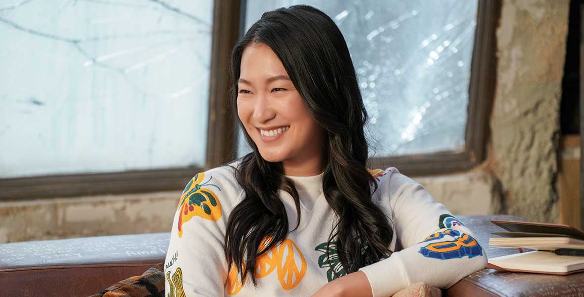 Actor Kara Wang smiles and sits on a brown, leather couch. She wears a white, long-sleeved sweatshirt emblazoned with blue and yellow butterflies, peace signs, and fruit, along with pants that feature yellow flowers and a bright pink stripe down the side of her leg. To her left is a small journal on the couch’s armrest.