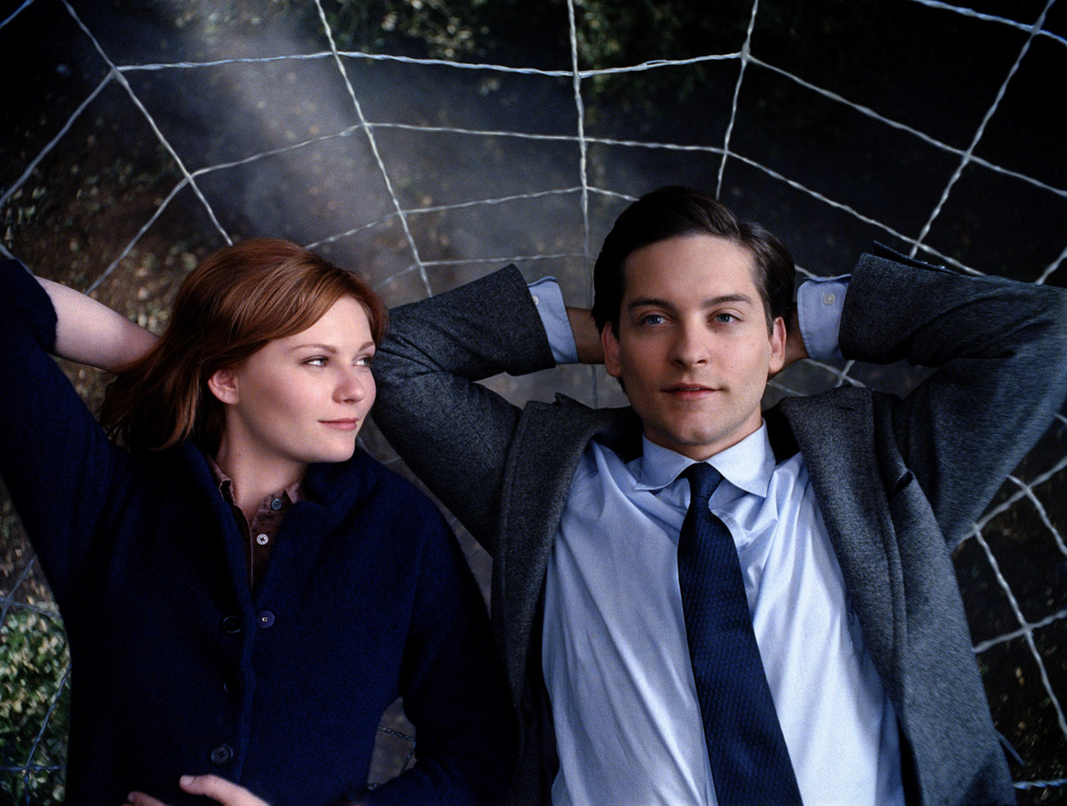 Peter Parker (Tobey Maguire) lays with his arms behind his head, as Mary Jane (Kirsten Dunst) looks at him with her right arm behind her head. She is wearing a black button up shirt. Peter is wearing a blue suit with a tie.