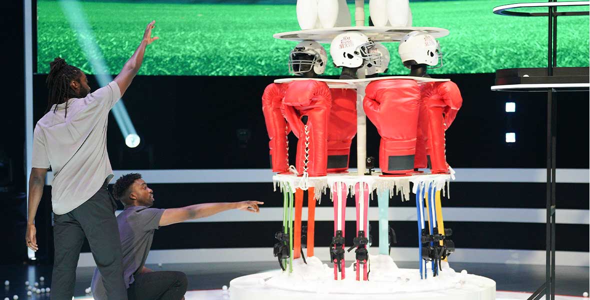 Two adult men face a giant tower featuring sport equipment-themed layers as the attempt to remove the necessary pieces on the game show The Final Show.