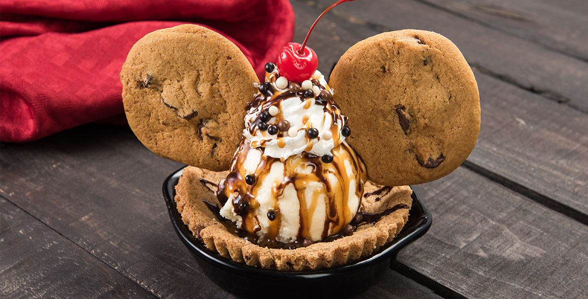 Vanilla ice cream is scooped on top of a cookie bowl with two cookies placed on either side of the scoop like Mickey ears. The sundae has caramel and chocolate drizzle with whipped cream, sprinkles, and a maraschino cherry on top.