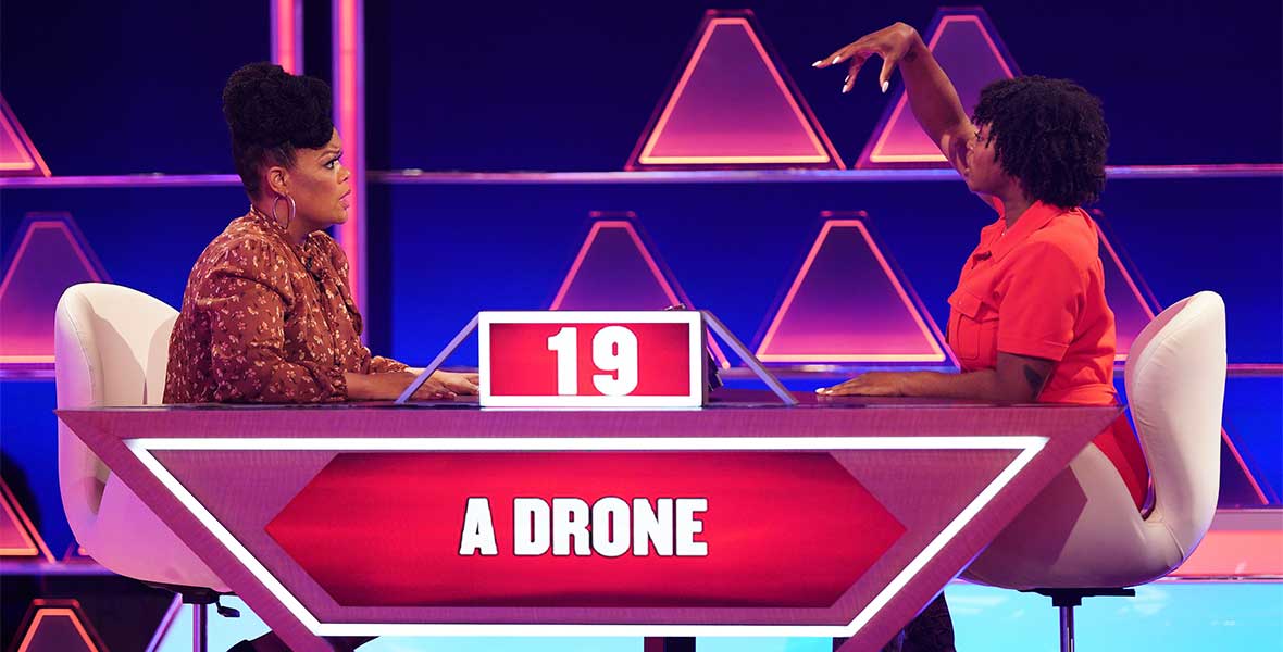 Actor Yvette Nicole Brown sits at a table displaying  the clue “a drone” and 19 seconds on the clock. Across from Brown is a female contestant with her right arm in the air acting out the clue. Behind them are bright lights forms in triangle shapes.