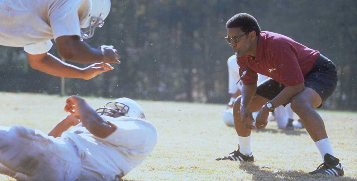 Actor Denzel Washington wears a maroon polo shirt and black shorts as he squats and leans forward. He looks at two football players tackling each other on a large green football field. Behind him are tall, leafy, green trees lining the field.