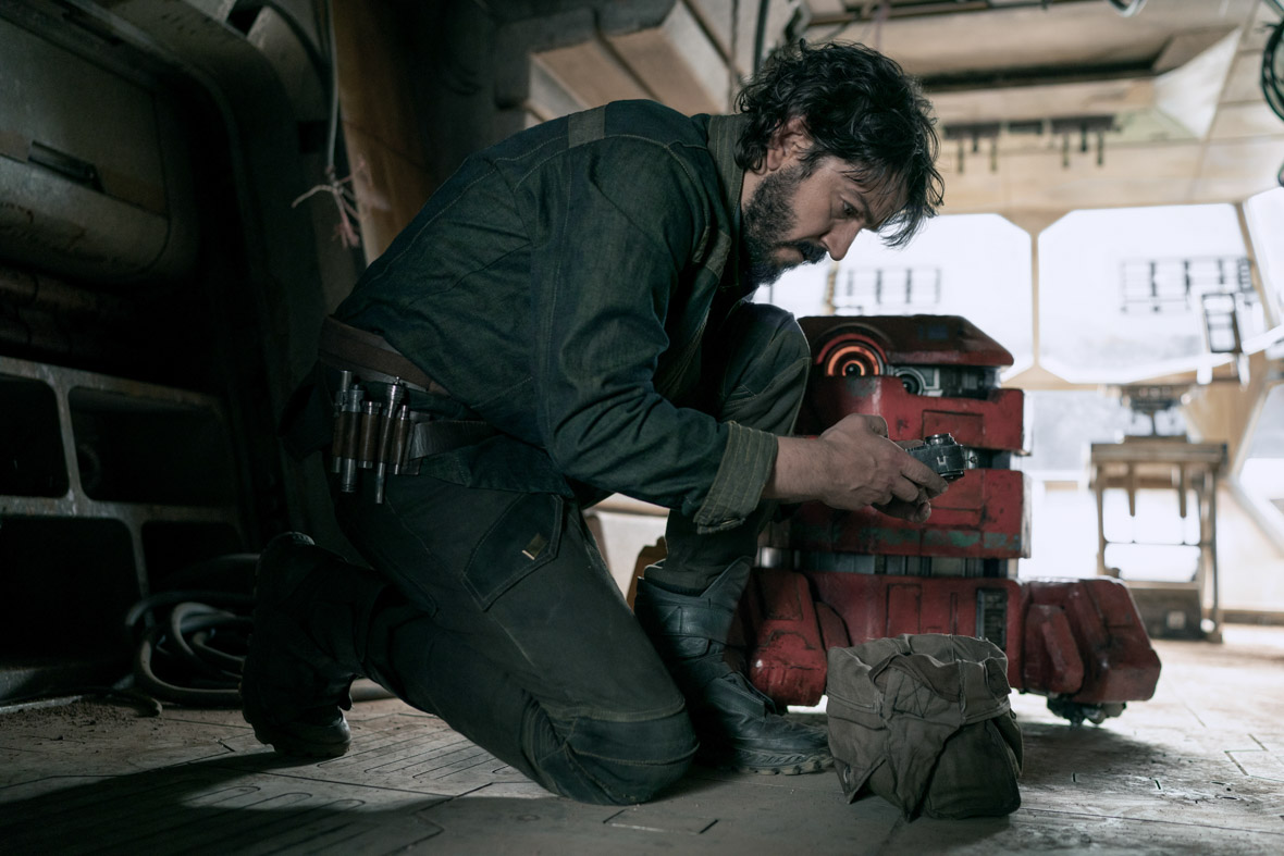 Cassian Andor, played by Diego Luna, wears denim coveralls. He is crouched down and tinkering with something, while the droid B2EMO stands behind him.