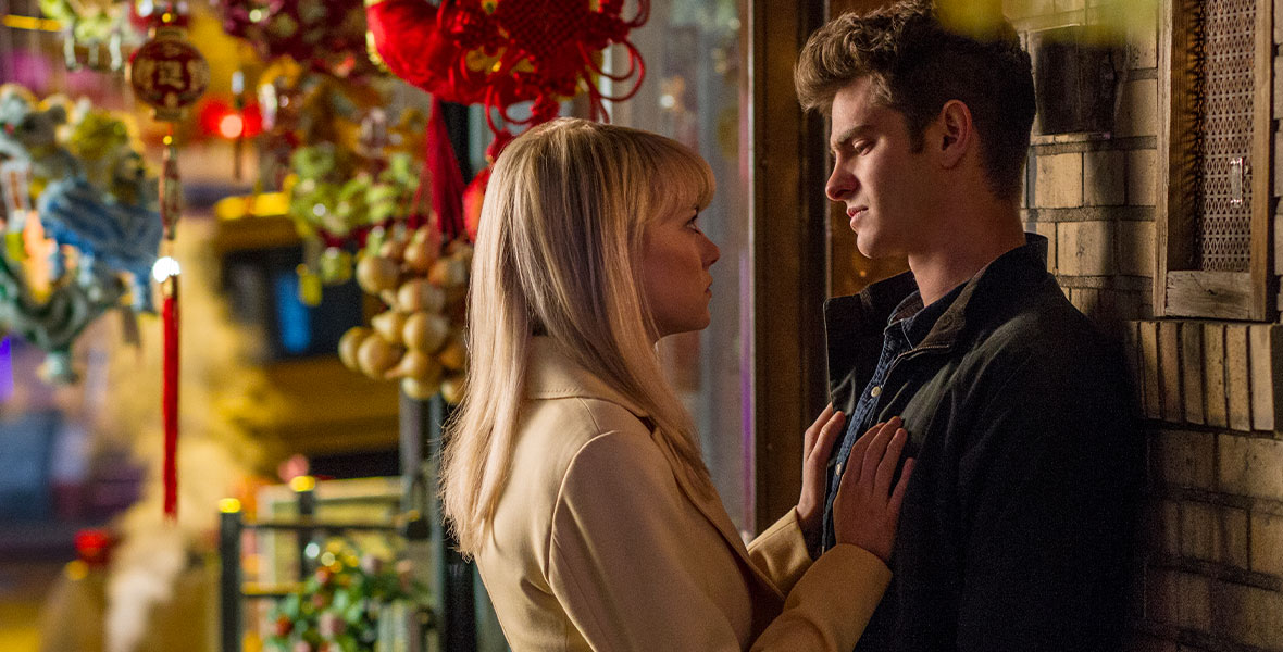 Peter Parker (Andrew Garfield) is leaning against a brick wall. Mary Jane (Emma Stone) has her hands on his chest as she looks up at him. She is blonde and wearing a tan peacoat, while Garfield is wearing a dark jacket.