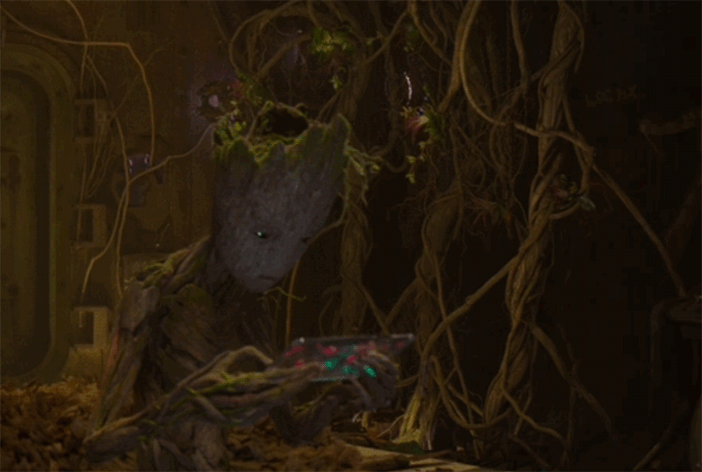 Teenage Groot from the post credits scene of Guardians of the Galaxy Vol.2. He is sitting on his bed in his room which is covered in large vines and moss. He looks disgruntled and plays a game on a handheld device.