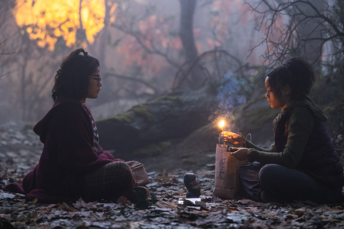 Izzy, played by Belissa Escobedo, and Becca, played by Whitney Peak, are seated opposite each other. They are in the woods holding a séance. Becca is about to light the Black Flame Candle.