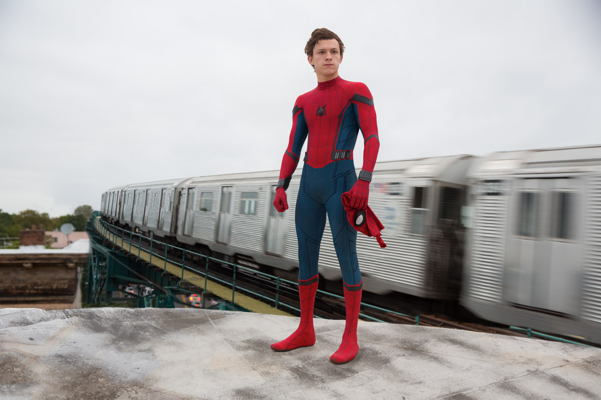 Peter Parker (Tom Holland) is in his Spider suit with his mask grasped in his left hand and a train passes behind him.