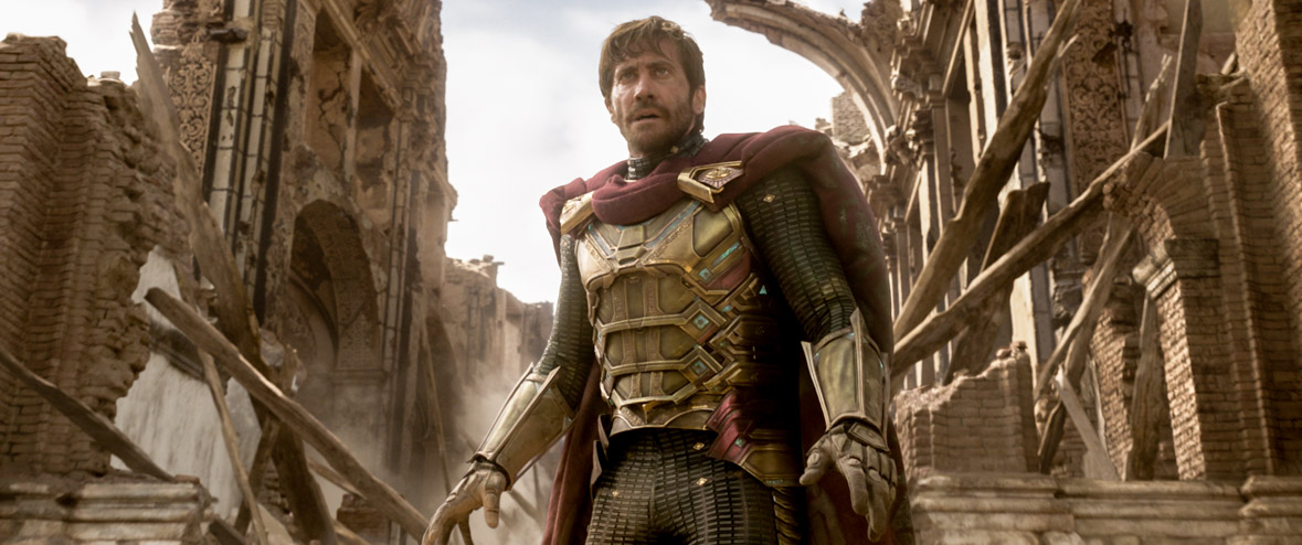 Mysterio (Jake Gyllenhaal) suited up in gold armor and a red cape amongst dusty buildings behind him.