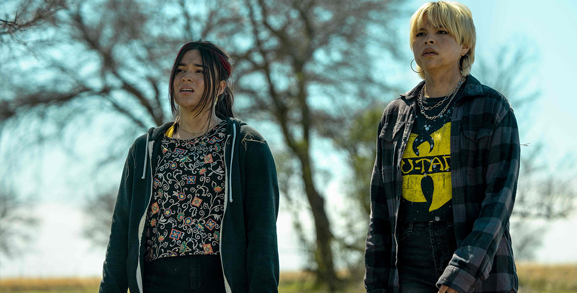 Actresses Devery Jack and Elva Guerra stand on a grassy hill with a bright blue sky and large tree in the background