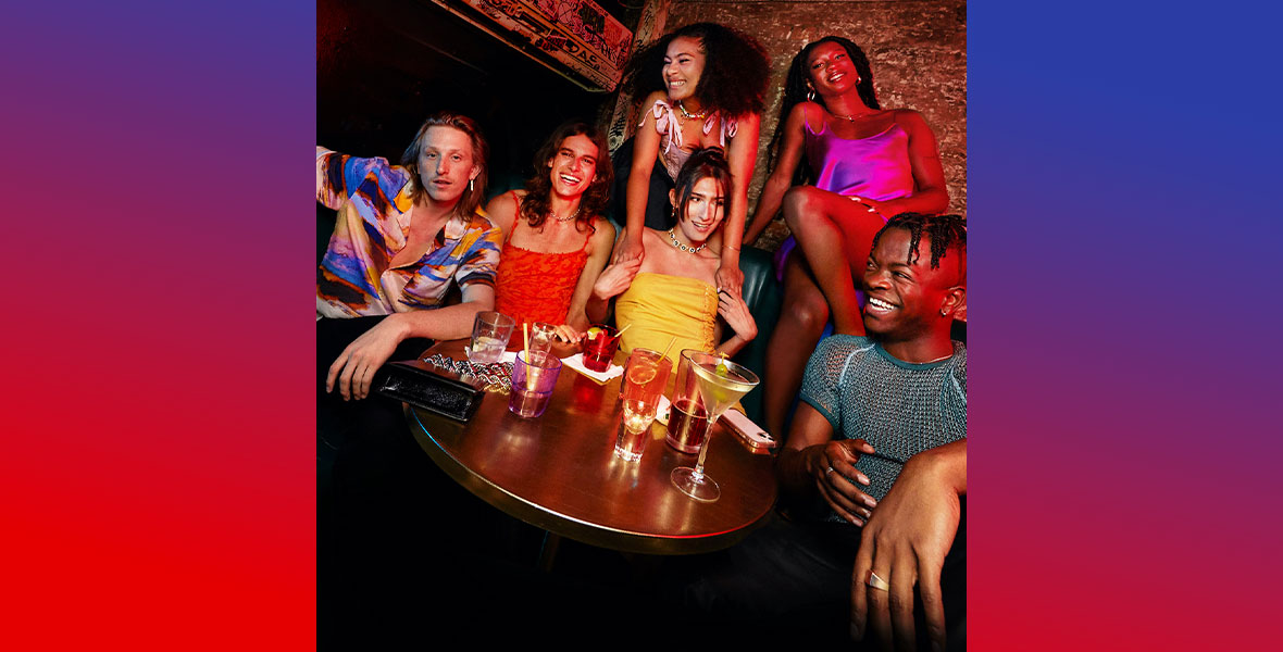 Left to Right: Ben Hard, Fernando Casablancas, Sophia Wilson, Claude Shwartz, Ebon Trower, andTaofeek Abijako. The cast of Freeform’s The Come Up wear trendy, colorful clothing as they sit around a circular table at a dive bar. They have cocktails in front of them, and the cast is laughing and smiling.