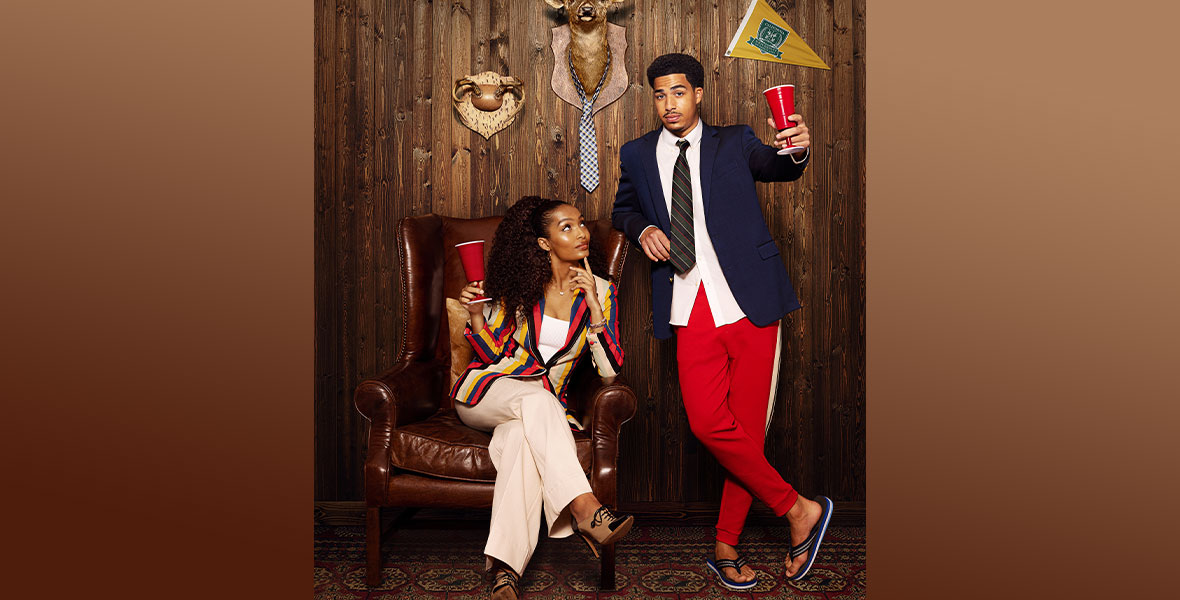 In the promotional poster for grown-ish Season 5, Yara Shahidi (left) is wearing a striped blazer and khaki pants. She is seated in a brown leather chair and holding a Solo cup, shaped like a chalice, and looking up at Marcus Scribner (right), who is wearing a striped tie, a white button-up shirt, a navy blue blazer, red track pants, and flip-flops. He, too, is holding a red Solo cup shaped like a chalice. Behind them is a wood paneled wall, featuring deer antlers, a deer’s head with a tie draped around its neck, and a pendant for California University.