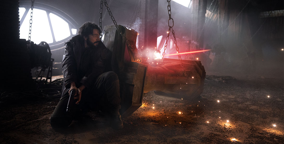 In a scene from Andor, actor Diego Luna crouches down on one knee and hides behind debris to avoid a blast. He is holding a blaster and is ready to make his next move.