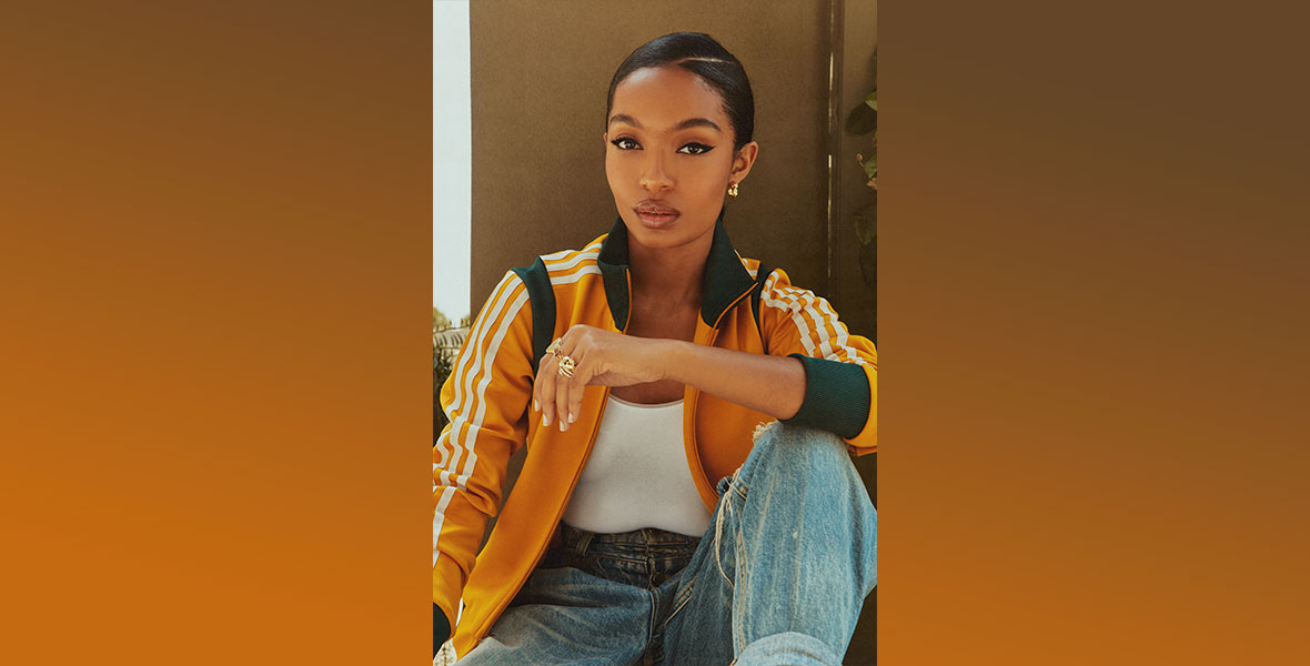 In this publicity shot of Yara Shahidi, she is sitting down with her left leg propped up and holding her left arm. She is wearing a gold track jacket with green trim and white stripes, a white tank top, and distressed denim jeans. Her hair is slicked back and she is wearing gold jewelry.