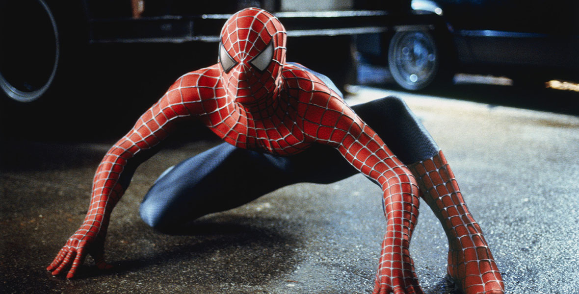 Spider-Man crouches down in his red and blue spidey suit, looking ready to pounce. His hands and knees are touching the asphalt.