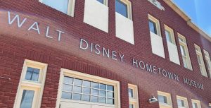 Close up photo of a large red brick building with silver letters stating: “Walt Disney Hometown Museum.” Above and below the letters lining the building are large white boarded windows. At the top of the building is a white plate stating “Marceline.”