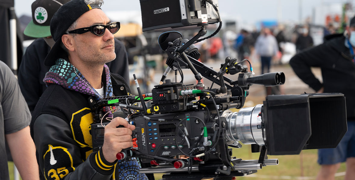 Taika Waititi wears a multicolored hoodie, sunglasses, and a backwards hat as he operates a camera on the set of Thor: Love and Thunder.