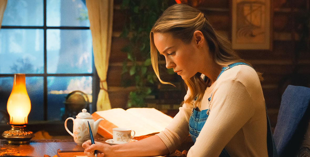 Brie Larson is seated at a desk and writing. Beside her is a teapot and a teacup, and a light. Brie’s hair is tucked behind her ears and she is wearing overalls.