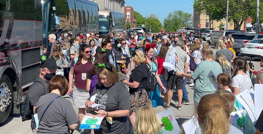 Crowd of guests getting off the black busses featured to the left of guests. Photo also features buildings of Marceline in the back against a bright blue sky.