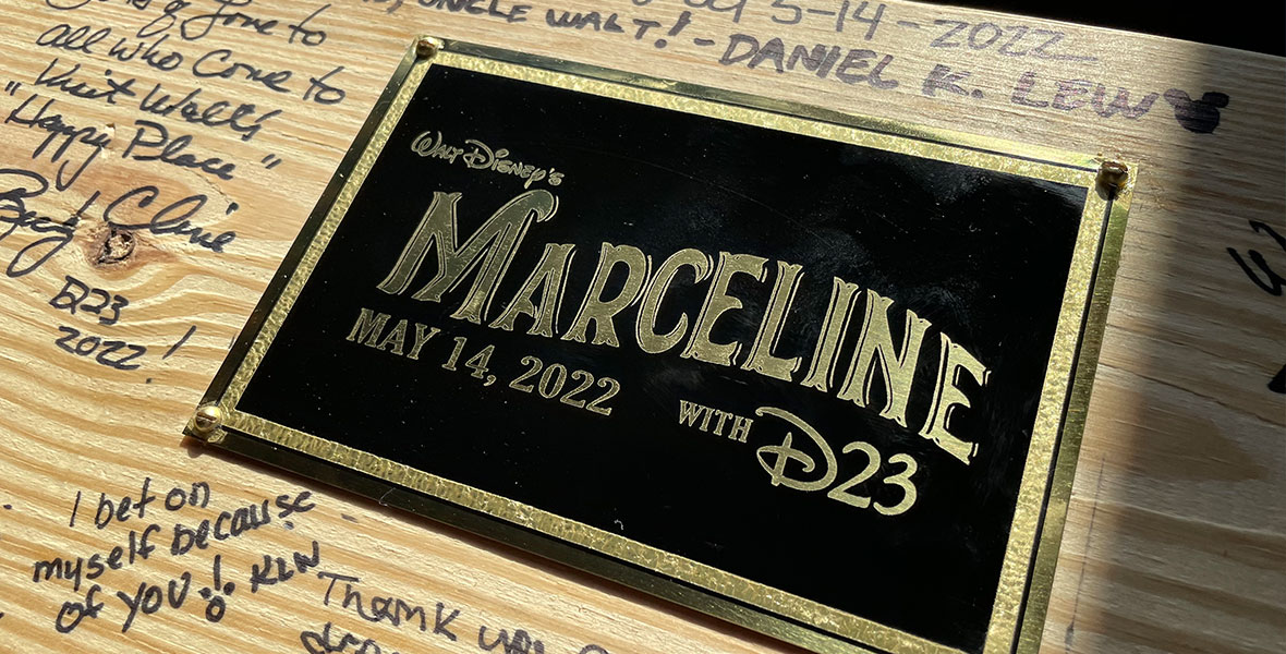A light wood background with a black and gold plaque featuring Walt Disney’s Marceline with D23 logo and the date of May 14, 2022, in gold lettering. Surrounding the plaque are signatures from D23 guests featuring names and small notes to Walt.