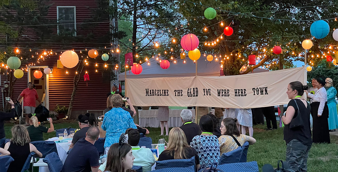 Many guests sitting and standing outdoors. The circular tables have white table clothes with bright pink and green flowers surrounded by folding chairs with blue checkered covers. Lights and lanterns are hanging above the tables and guests in green, pink, red, and yellow. Behind the tables to the right is a large, rectangular white fabric banner with black text reading “Marceline The Glad You Were Here Town.” Several large green trees and a red barn can be seen in the back left of the photo.