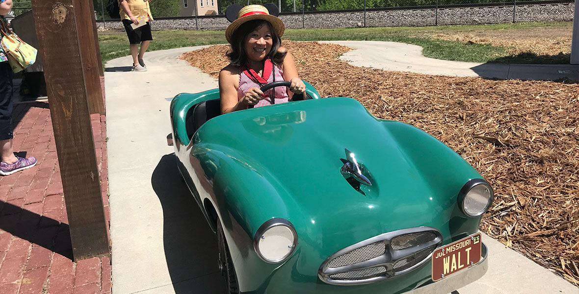 A guest sitting in an emerald green retro car with silver rim and white headlights with brown license plate on the front right of the car featuring Walt in big letters, Jul in the top left and Missouri above Walt. The guest is smiling and has short dark hair, a red and white striped tank top and a woven sun hat with red band and black Mickey ears on either side of the hat. The car is on the sidewalk with brick flooring on the left with brown beams reaching toward the ceiling. On the right side of the car are wood chips and green grass. The background features green trees and a blue sky.