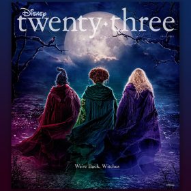 The front cover of Disney twenty-three, featuring the publication logo above the backs of Sarah, Winnifred, and Sarah Sanderson, all wearing cloaks and standing on a wooded hill overlooking a town at night. The text below them states, “We’re back, witches.”