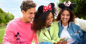 Three people gathered together looking at a phone. The person on the left wears a bright red Mickey Mouse shirt, while the two people beside him wear Minnie Mouse ear headbands. Everyone is smiling as they look at something on the phone.